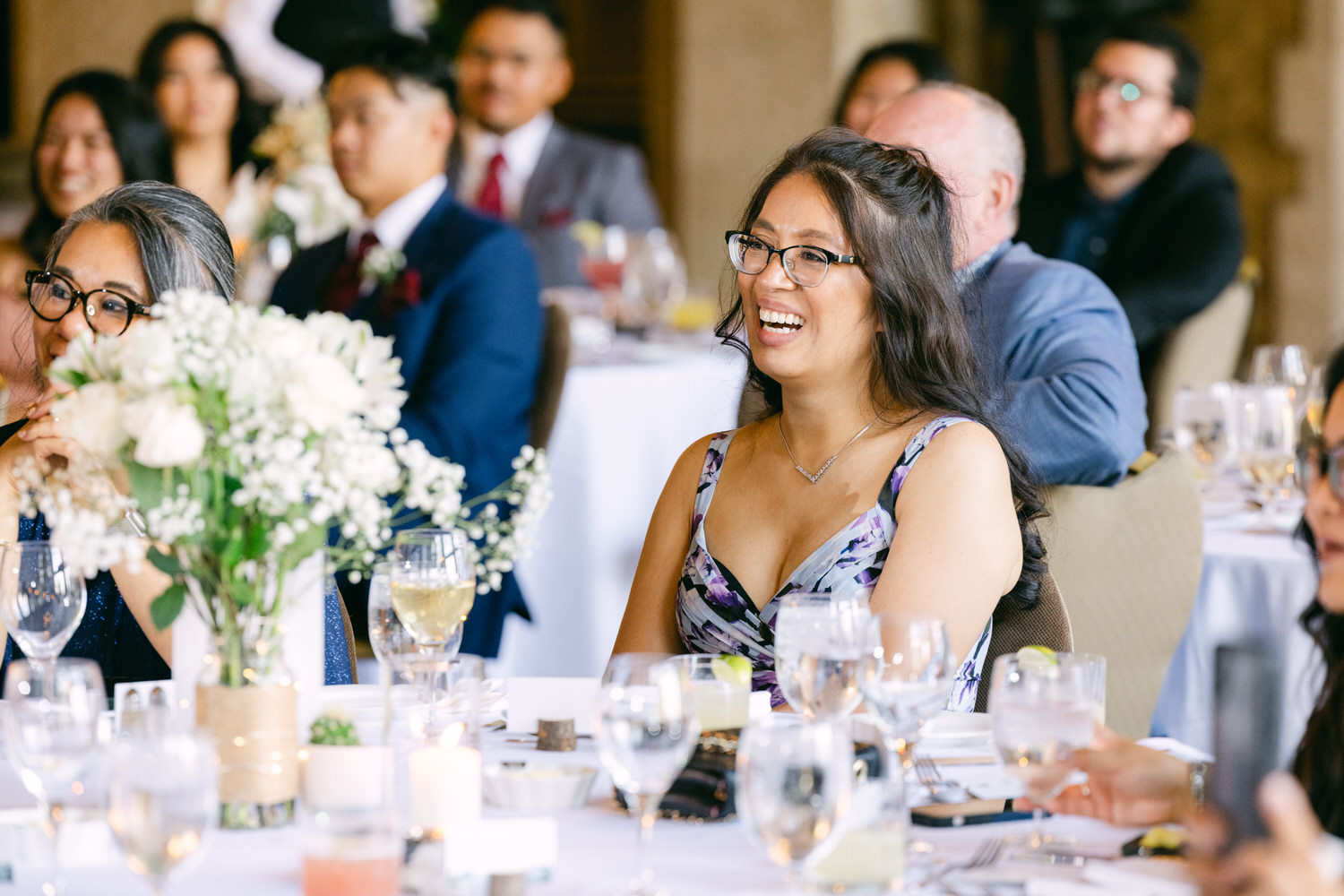 A smiling woman enjoying a moment at a festive gathering, surrounded by guests and floral arrangements at elegantly set tables.