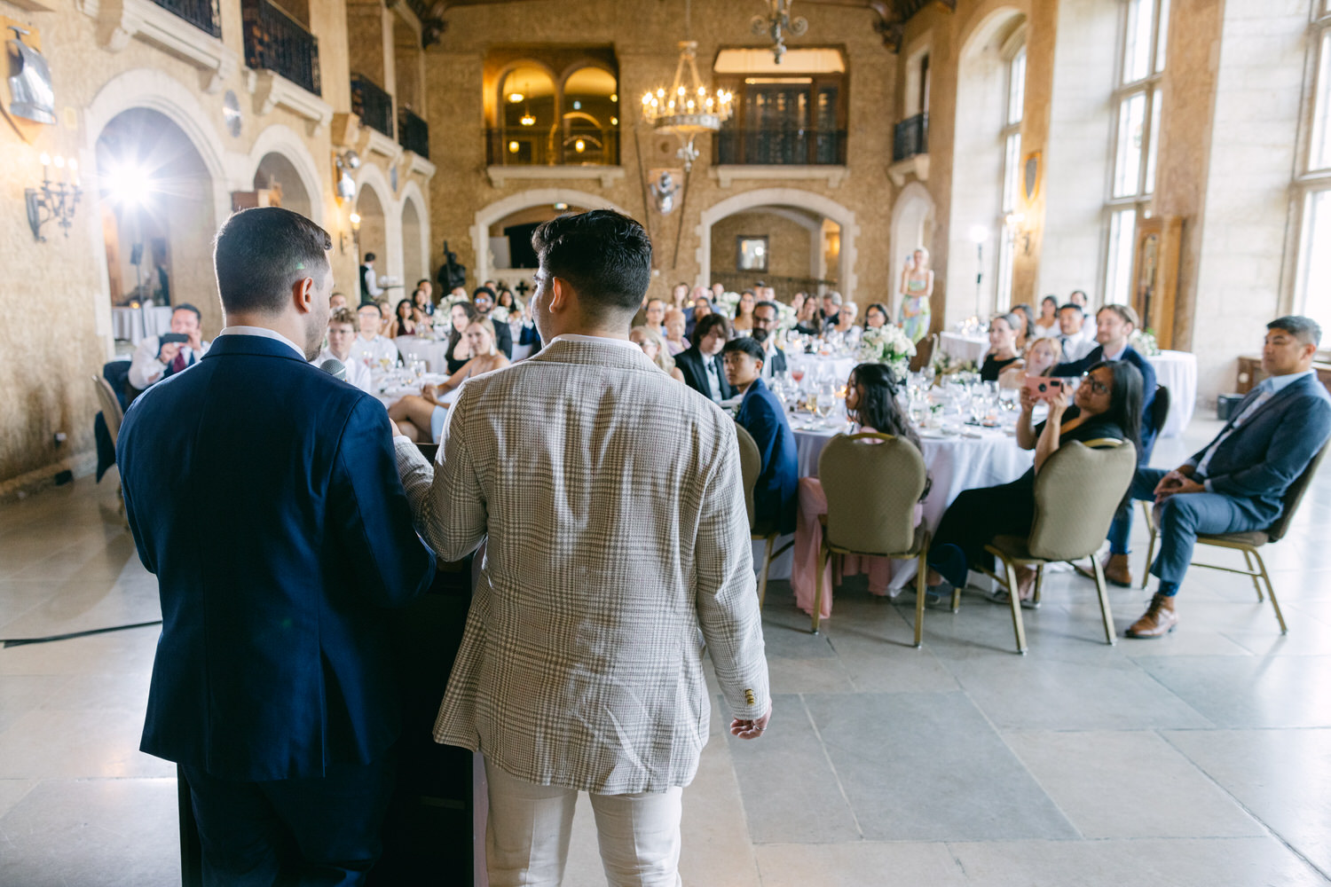 Two speakers address a wedding crowd in an elegant venue, with guests seated at tables enjoying the moment.