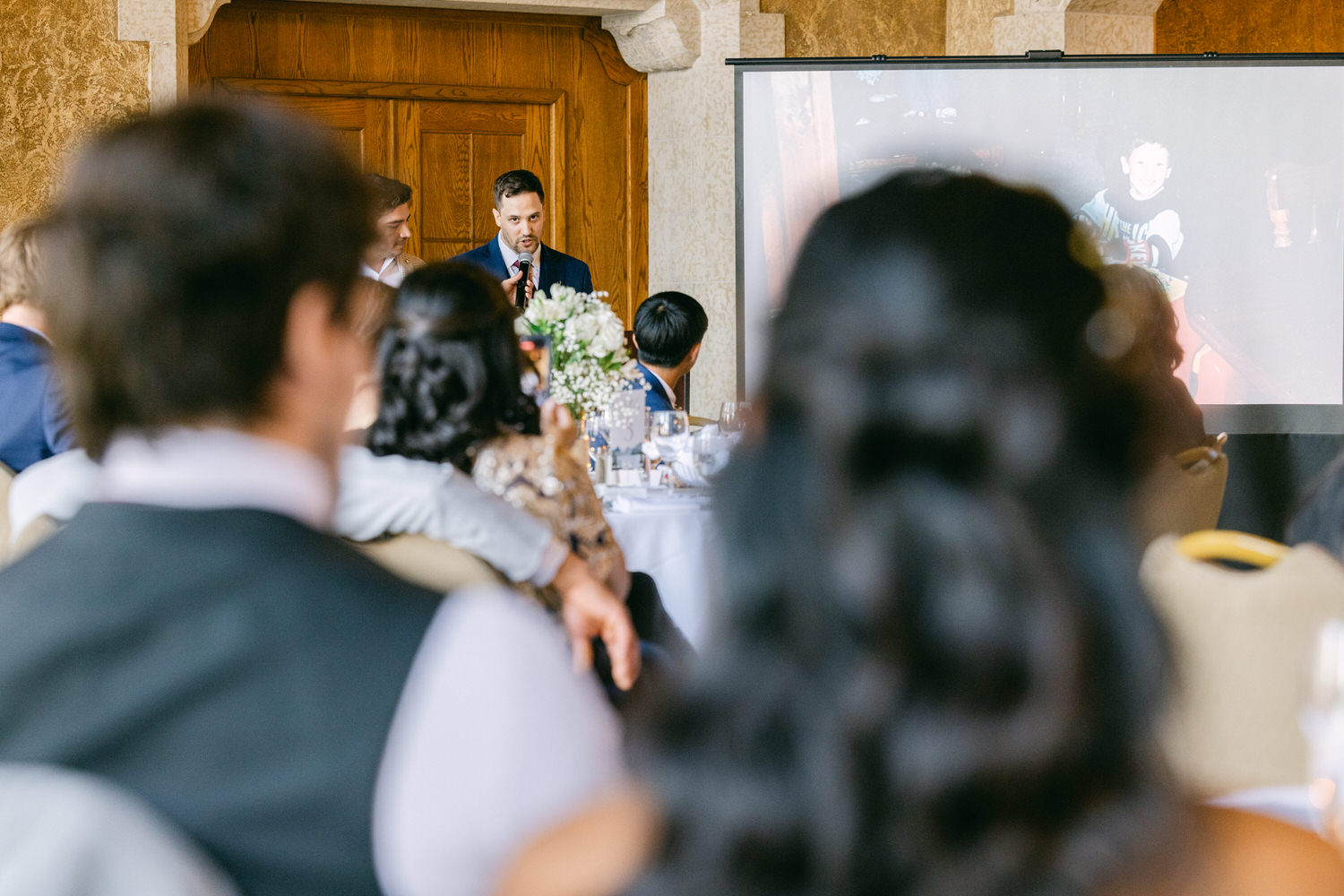 A man speaking at a wedding while guests listen, with a presentation screen showing a childhood photo in the background.