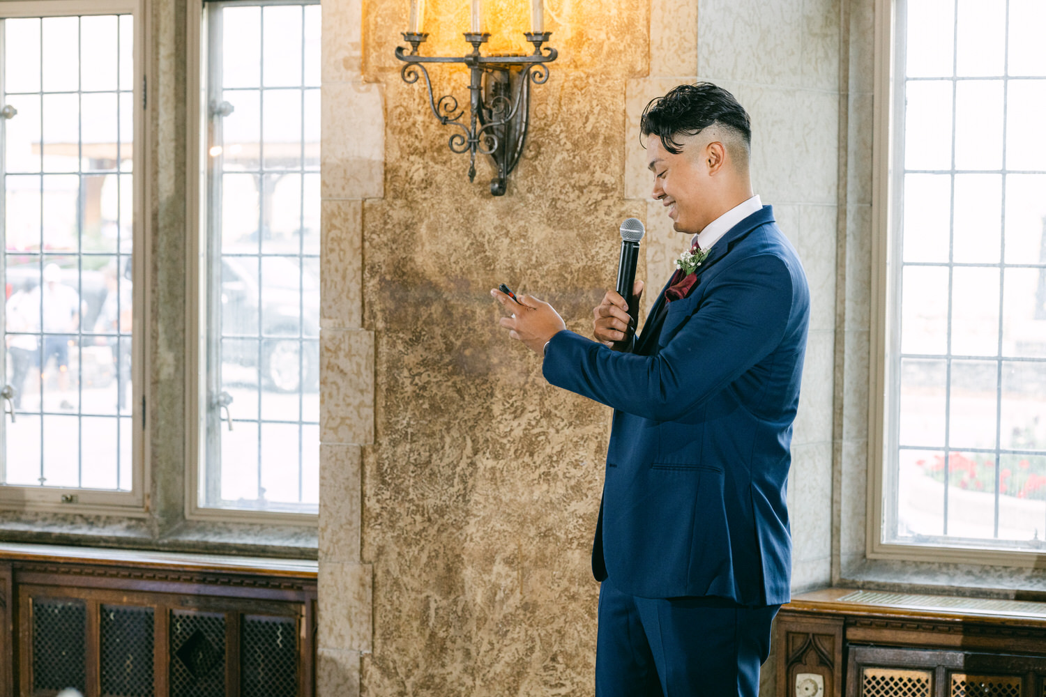 A man wearing a blue suit holds a microphone and looks at his phone while speaking at a wedding reception, with large windows in the background allowing natural light.