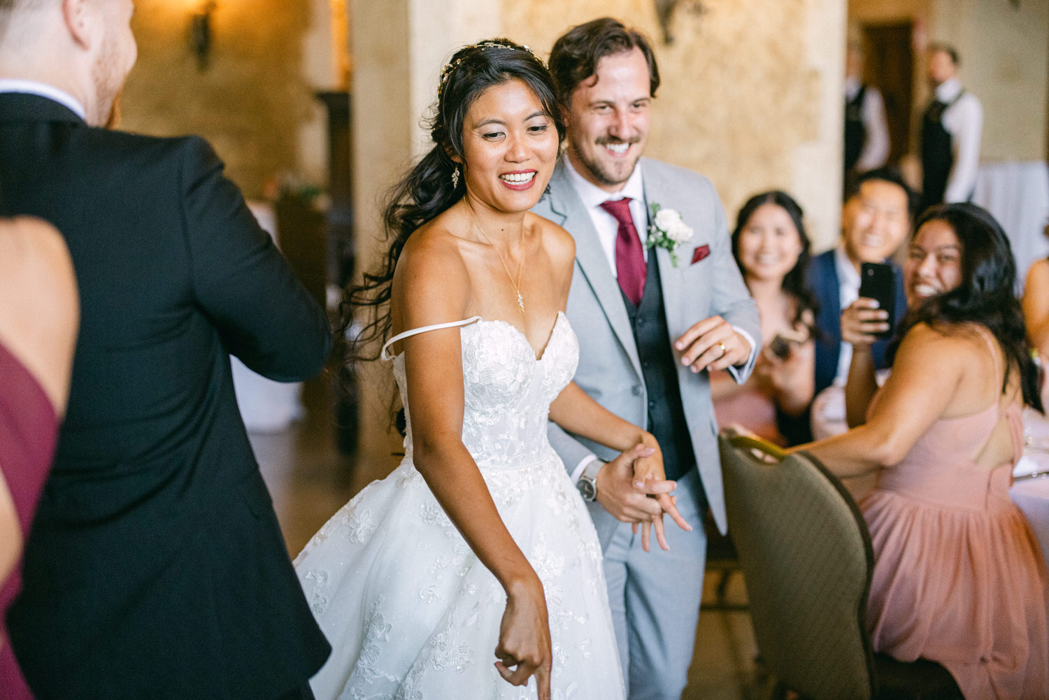 A bride in a beautiful gown dances with her groom during a joyful wedding reception, surrounded by smiling guests.