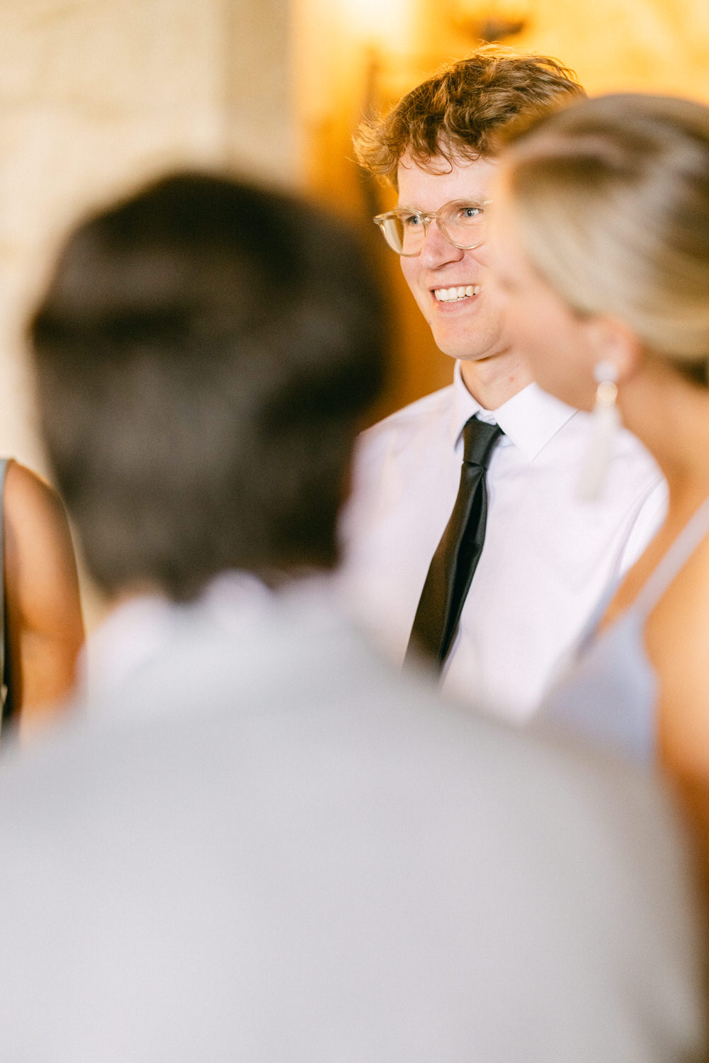 A smiling man in formal attire engages in conversation with friends, set against a softly blurred background.