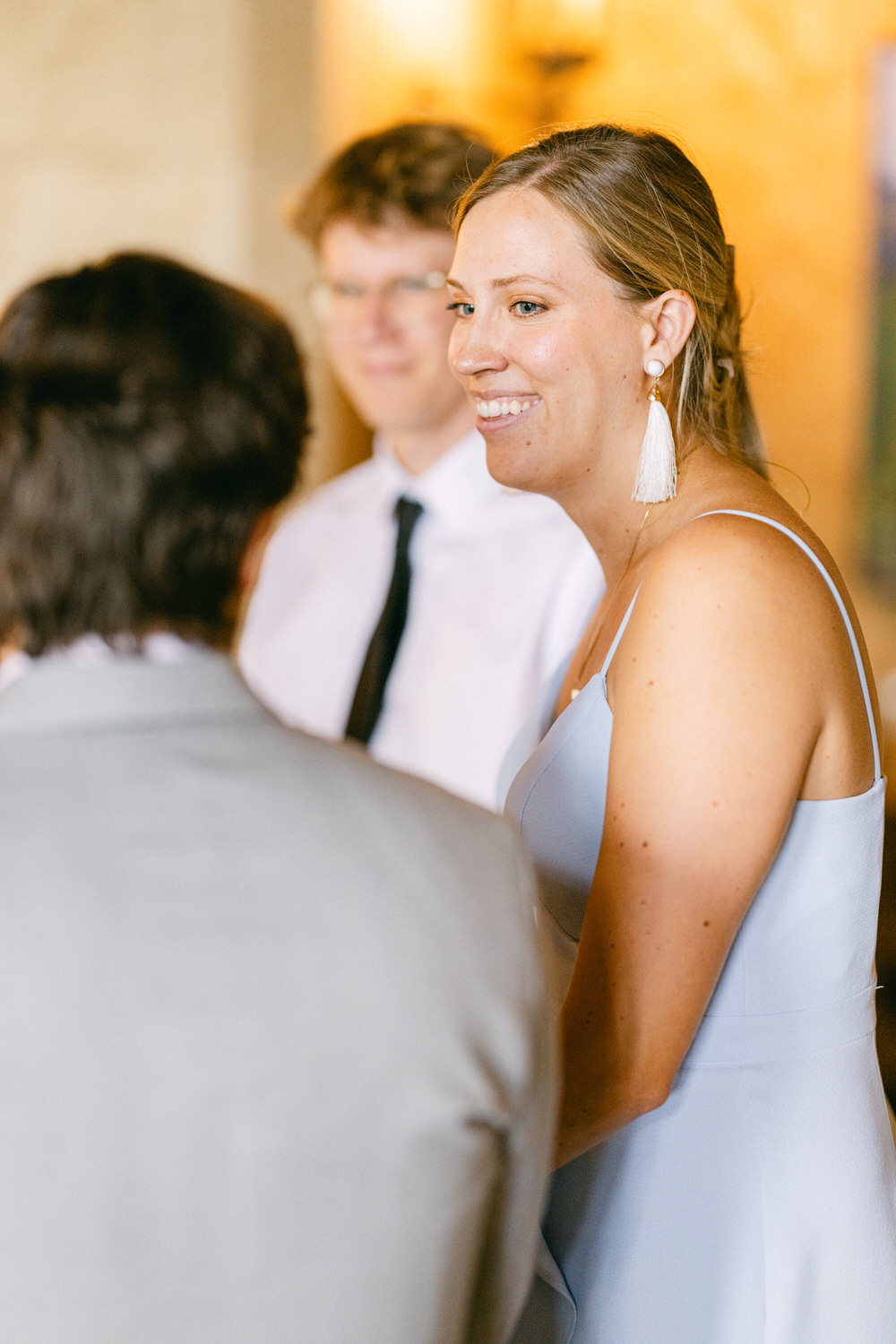 A woman in a light blue dress smiles while engaging in conversation with two individuals, set in a softly lit indoor space.