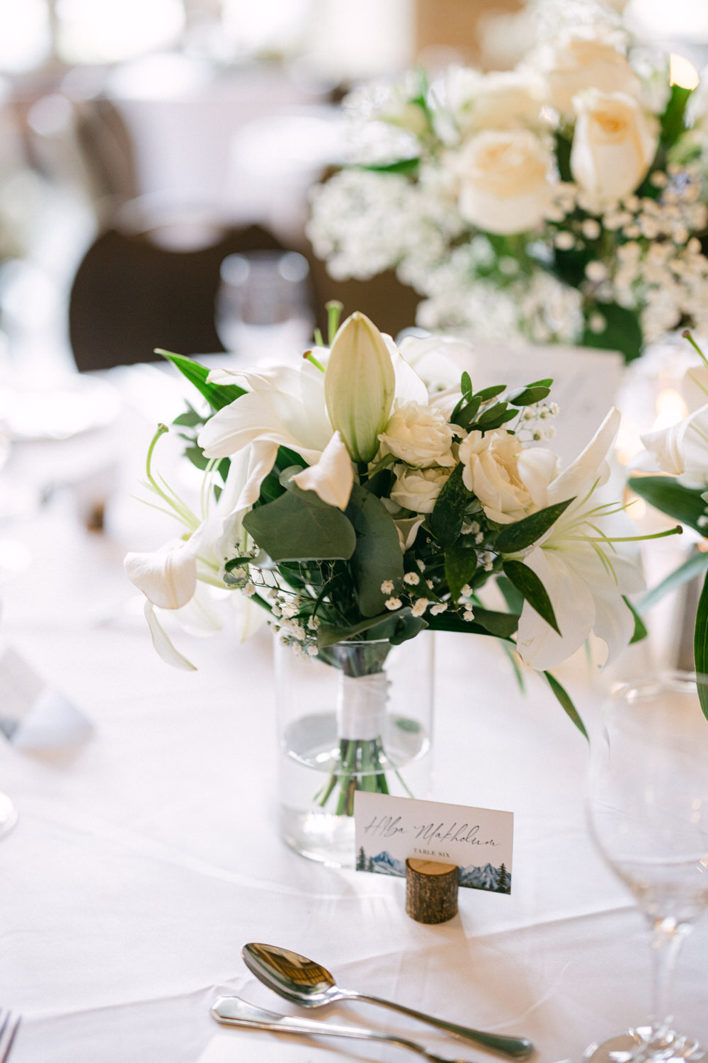 A beautifully arranged bouquet of white lilies and roses in a glass vase, accompanied by a place card on a wooden holder, set on a dining table.