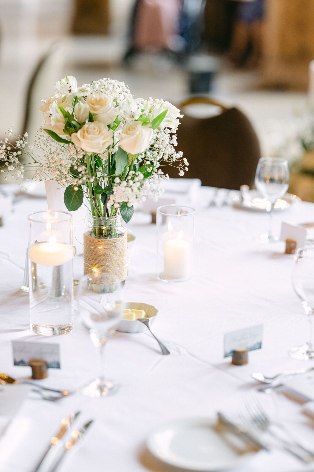 A beautifully arranged table featuring a floral centerpiece of white roses and baby's breath, surrounded by candles, glassware, and silverware on a white tablecloth.