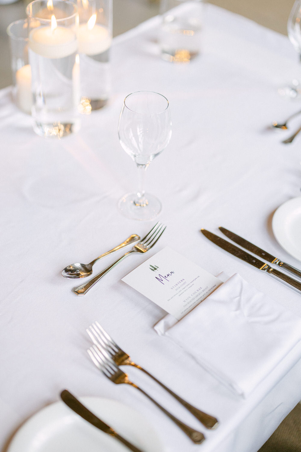 A beautifully arranged dining table featuring sparkling glassware, cutlery, and a menu card on a crisp white tablecloth, complemented by floating candles in clear vases.