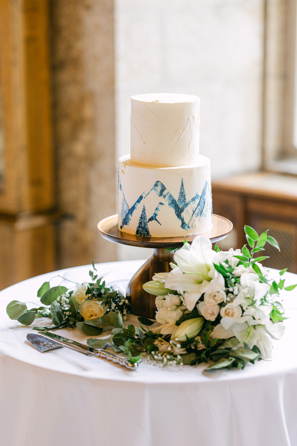 A beautifully decorated three-tier wedding cake with mountain and pine tree designs, accompanied by a bouquet of white flowers and greenery on a wooden pedestal.