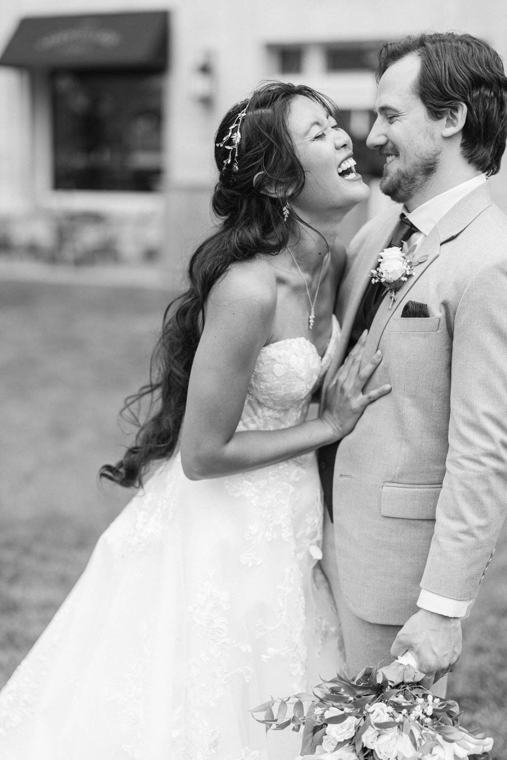 A couple sharing a heartfelt laugh in a romantic embrace, surrounded by greenery, showcasing love and happiness on their special day.