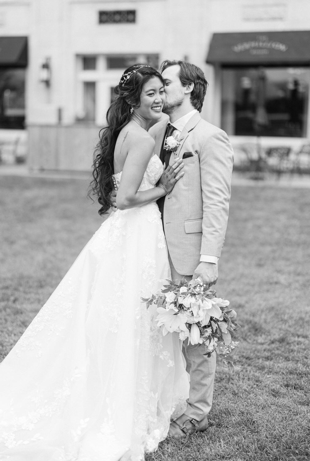 Bride and groom share a joyful embrace outdoors, with the bride in a stunning white gown and the groom in a light gray suit, holding a bouquet of flowers.