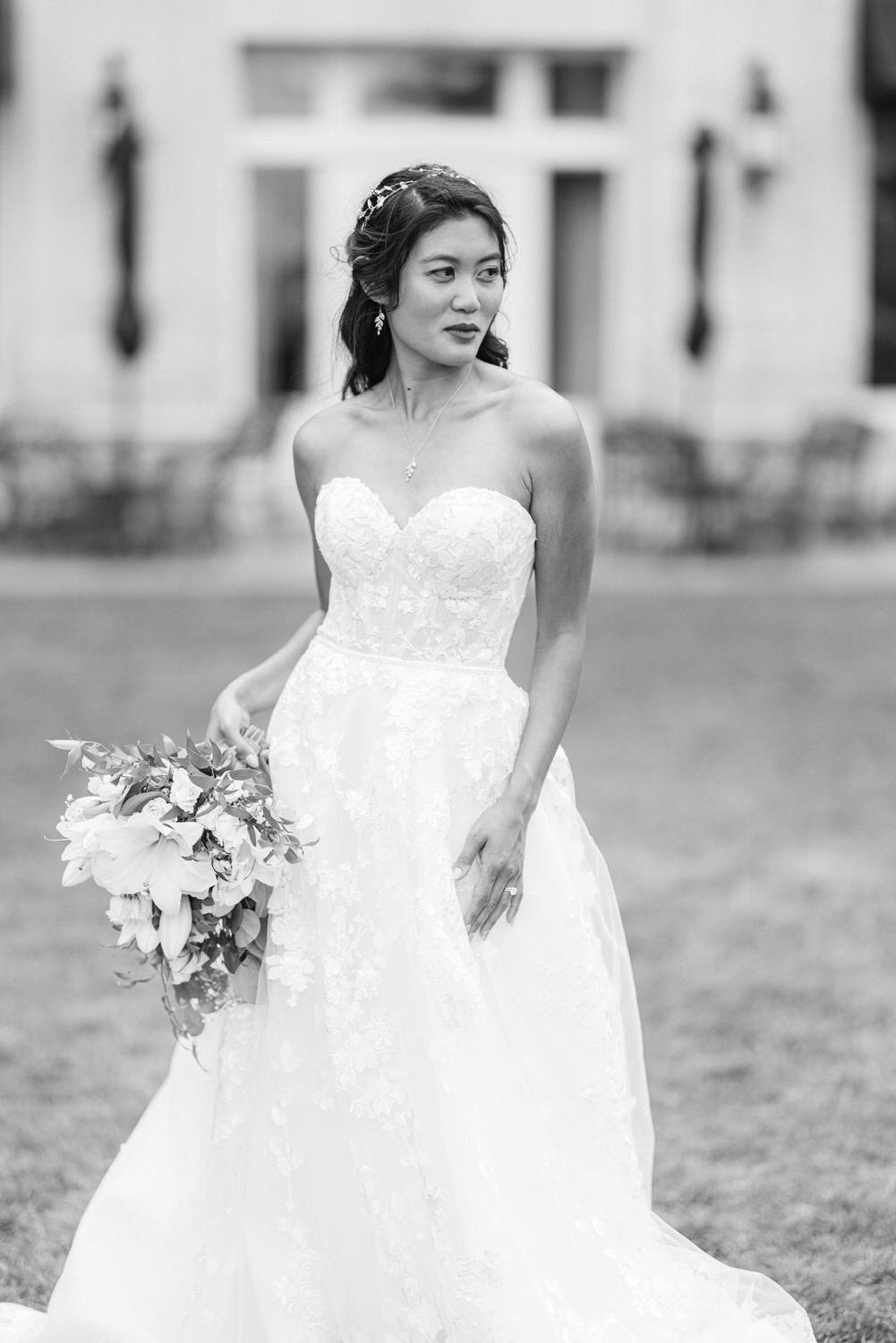 A bride poses outdoors in a beautiful lace wedding dress, holding a bouquet of flowers, with a serene expression.