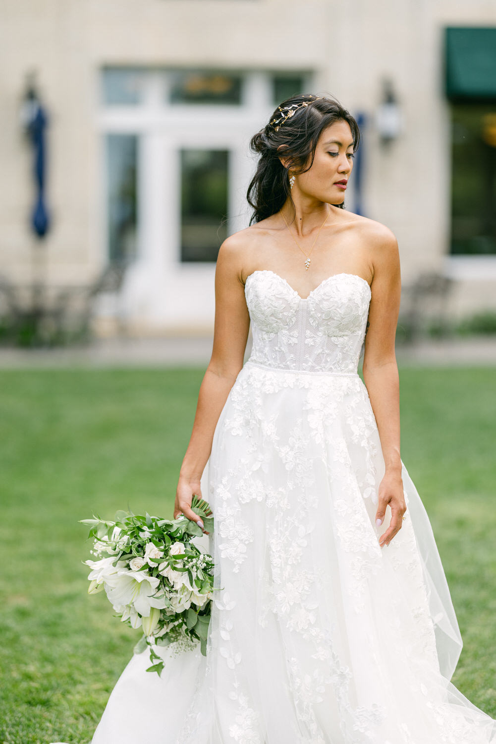 A bride wearing a strapless floral wedding gown holds a bouquet while gazing thoughtfully outdoors.