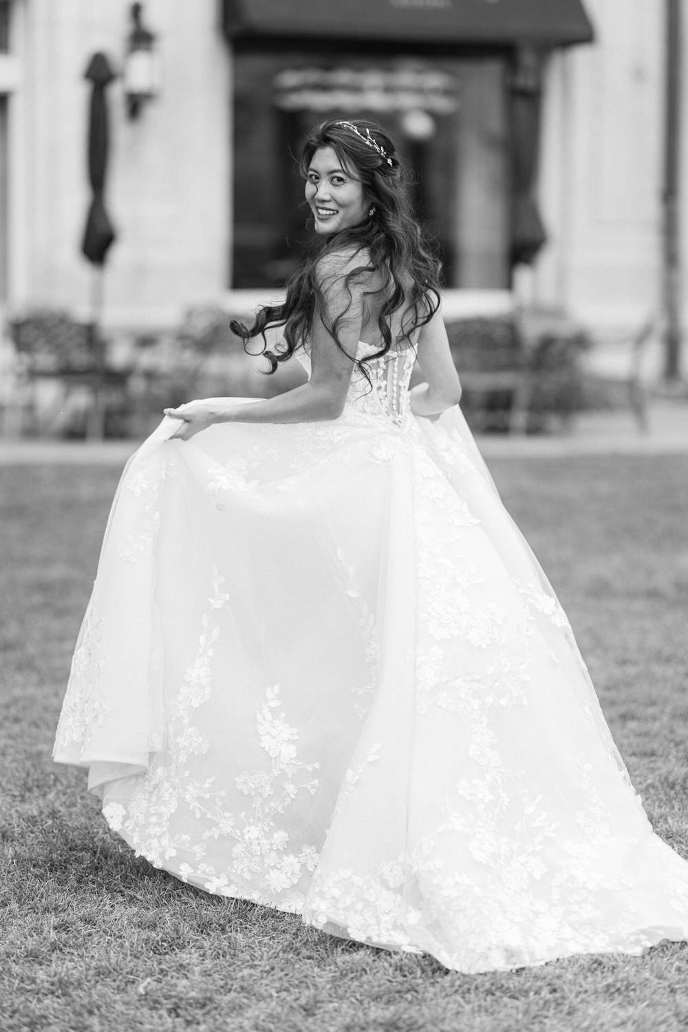 A joyful bride twirls in a flowing white gown adorned with floral lace, smiling over her shoulder in a picturesque outdoor setting.