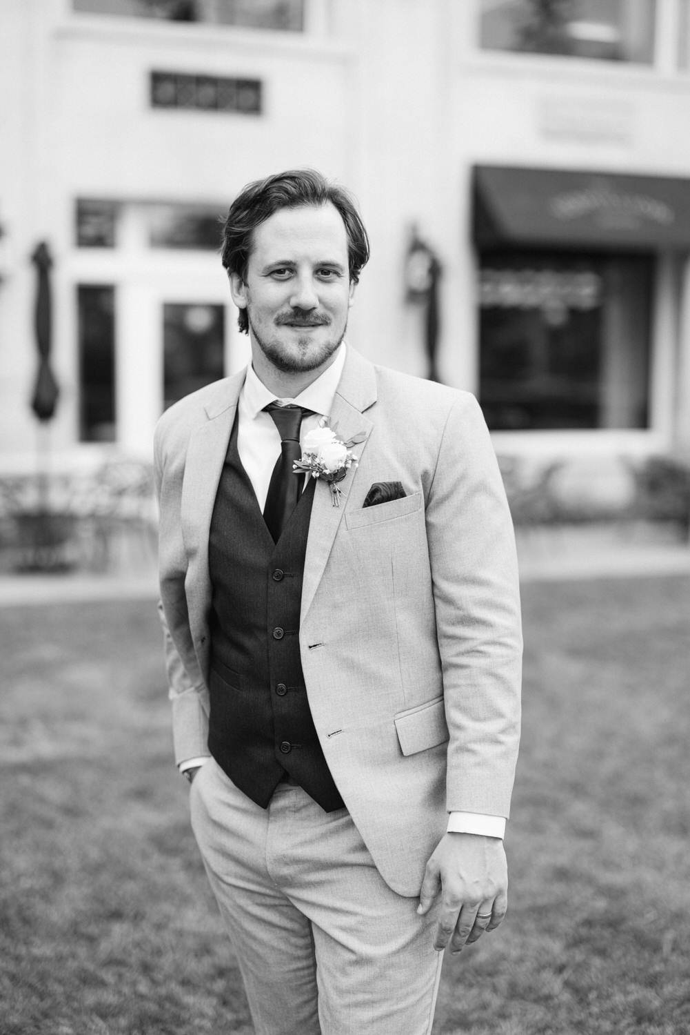 A well-dressed man in a suit and vest stands outdoors, smiling confidently with a boutonniere on his lapel.