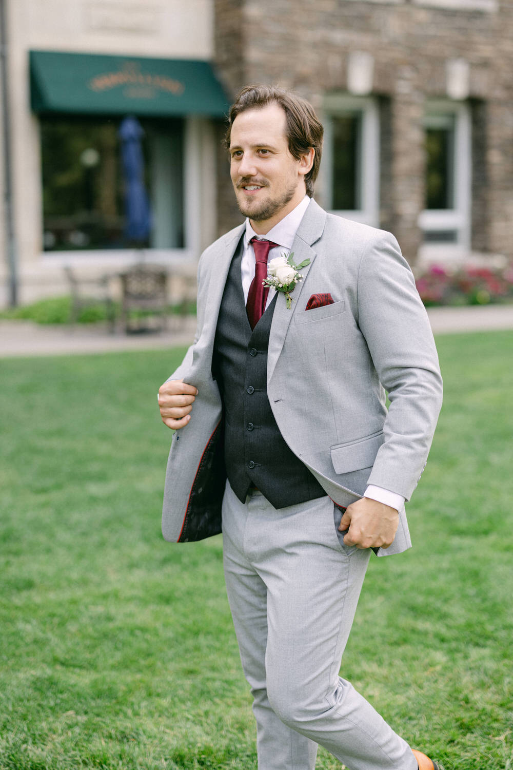 A well-dressed man in a light grey suit and burgundy tie walks confidently on a lawn, showcasing a white boutonnière and a stylish pocket square.