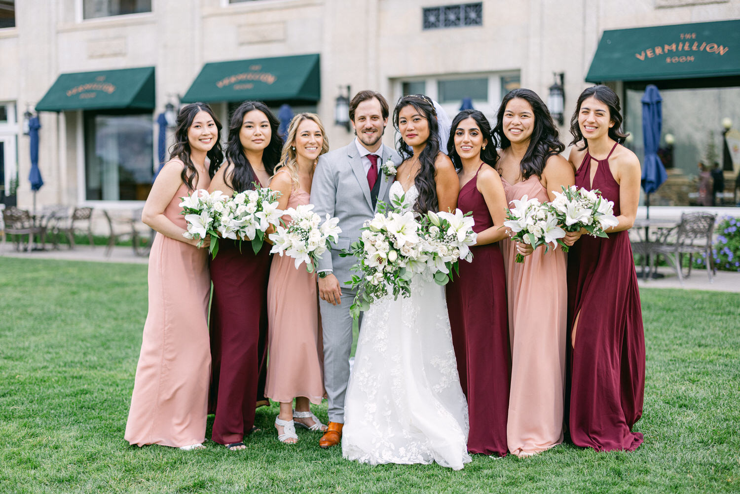 A smiling couple poses with their bridal party, featuring women in varying shades of pink and burgundy dresses, all holding white floral bouquets in a lush outdoor setting.