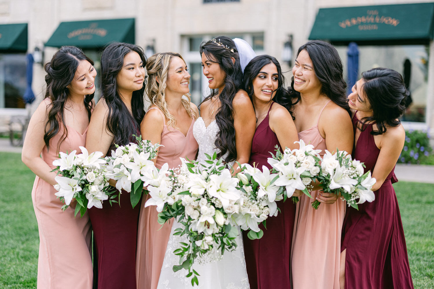 A bride surrounded by her six bridesmaids, all smiling and holding beautiful floral bouquets, standing in a lush outdoor setting.