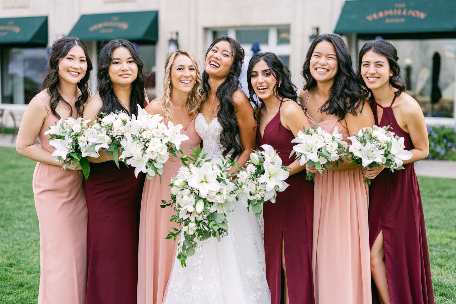 A joyful group of bridesmaids wearing a mix of pink and burgundy dresses, holding elegant bouquets of white lilies and greenery, posing together outdoors.