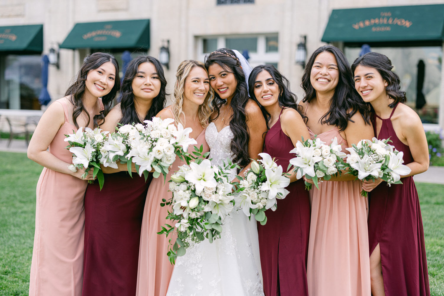 A group of seven bridesmaids, wearing elegant dresses in pink and burgundy, gather around the bride, who is in a white gown, each holding bouquets of white flowers in a lush green outdoor setting.