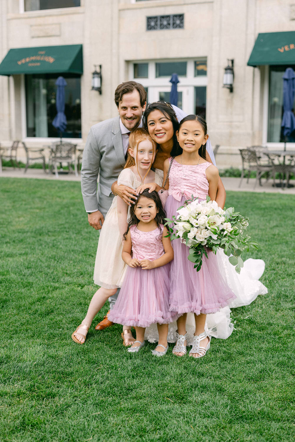 A joyful family poses together outdoors in wedding attire, featuring a bride with a bouquet and three children in matching dresses, surrounded by lush green grass and a charming venue backdrop.