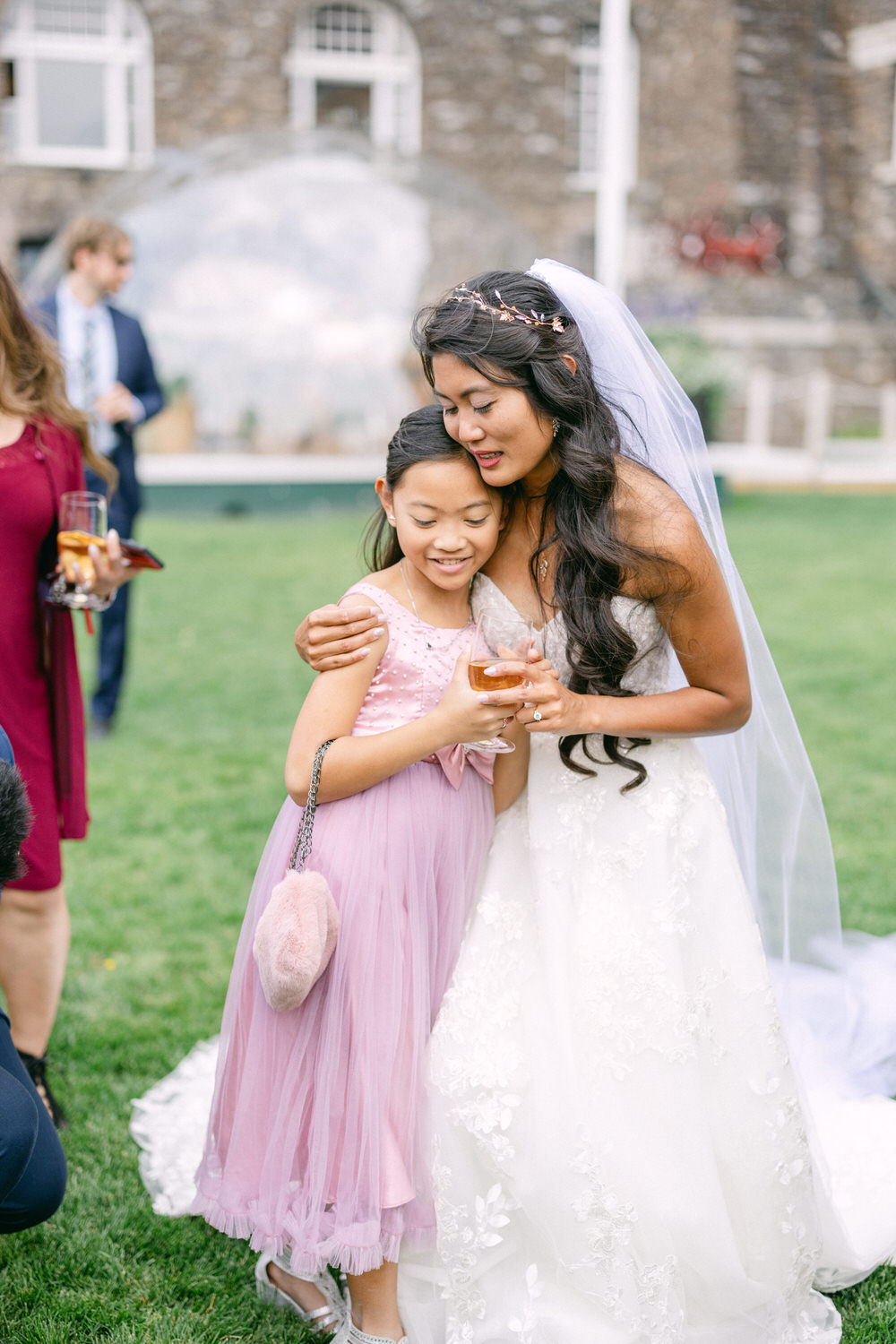 A bride embraces a young girl in a pink dress, both smiling, surrounded by wedding guests in a lush outdoor setting.