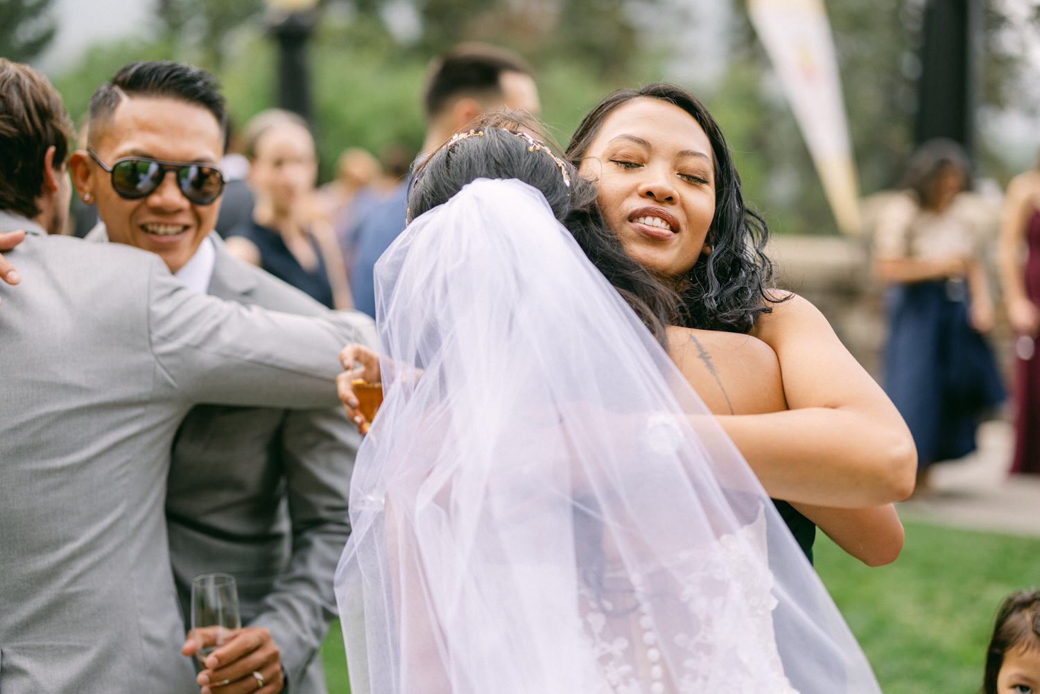 Two women joyfully hugging at a wedding, surrounded by guests in a lush outdoor setting.