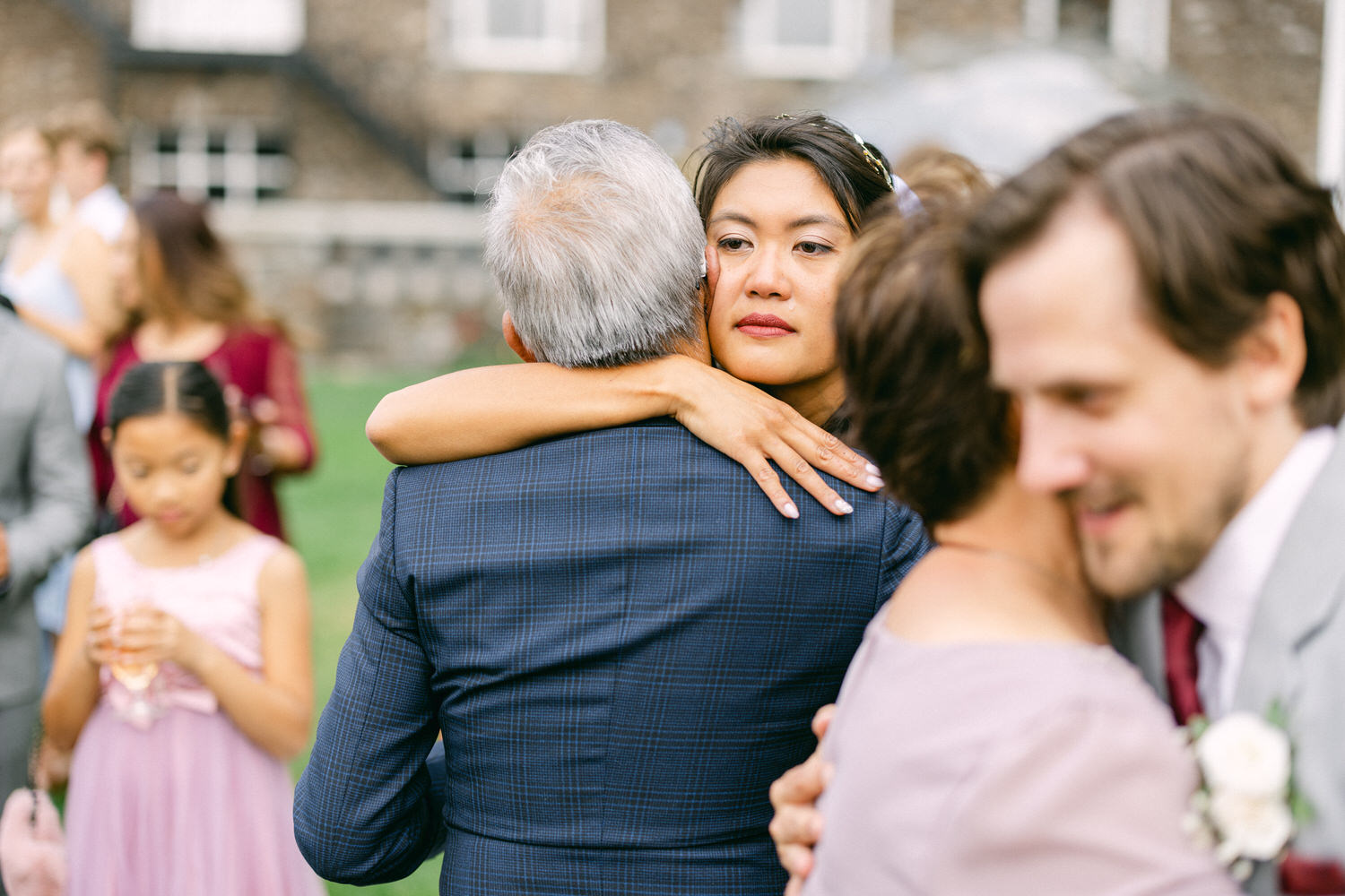 A woman hugs a man with gray hair while gazing thoughtfully, surrounded by guests enjoying a festive outdoor event.