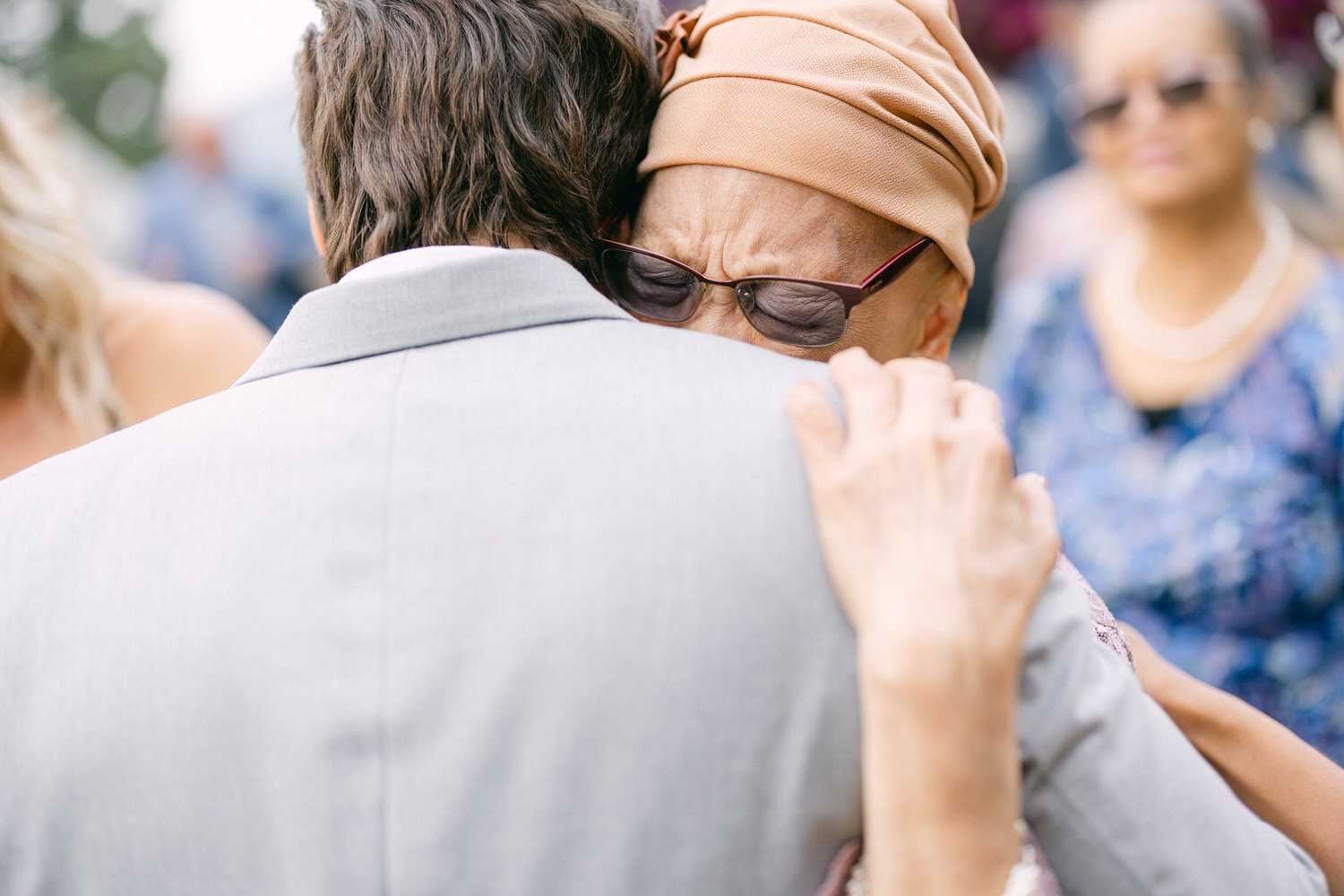 A man in a light gray suit hugs an elderly woman wearing a headscarf and glasses, conveying deep emotion amidst a joyful outdoor gathering.