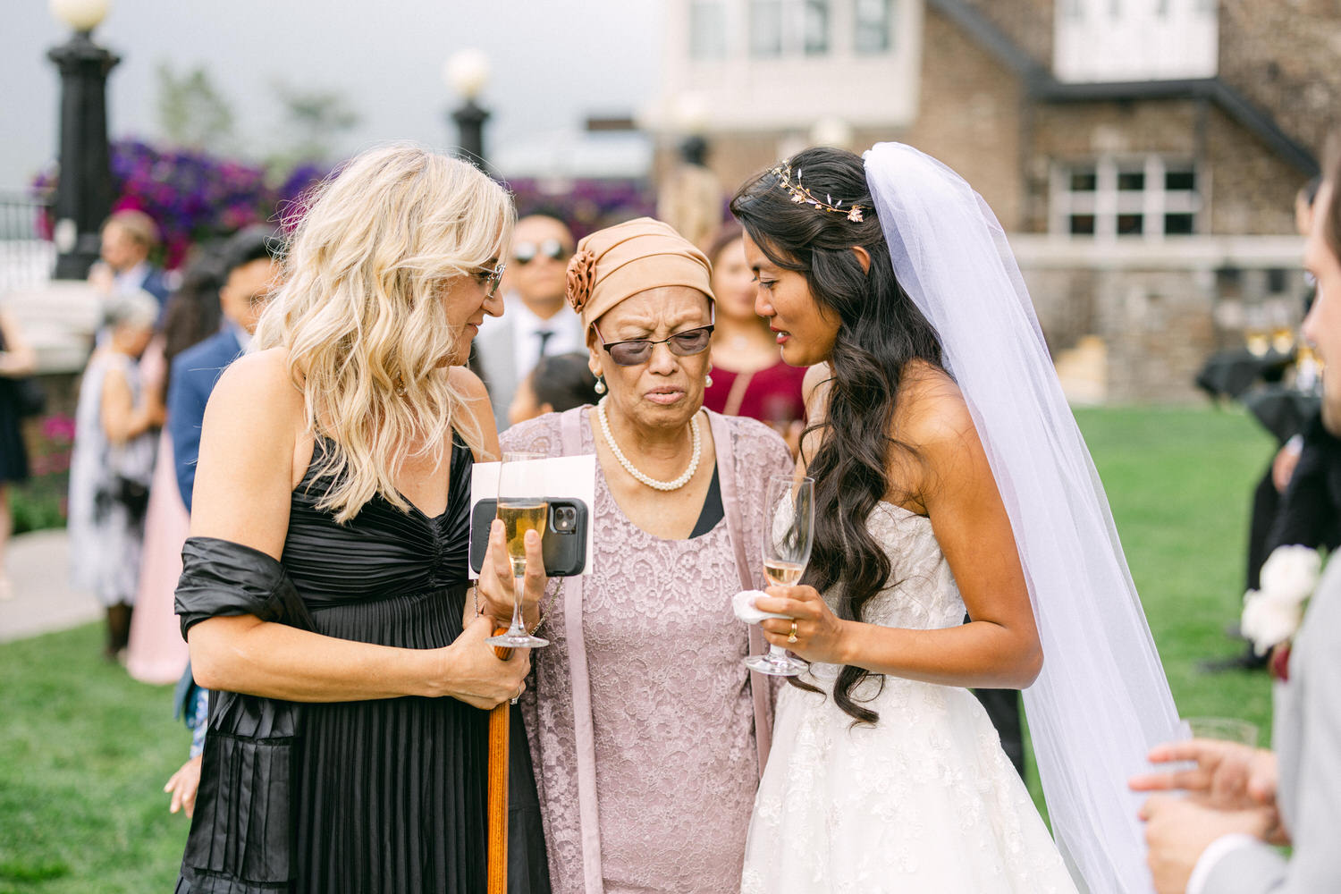 A bride in a strapless gown interacts joyfully with a grandmother and a friend, surrounded by wedding guests in a beautiful outdoor setting.