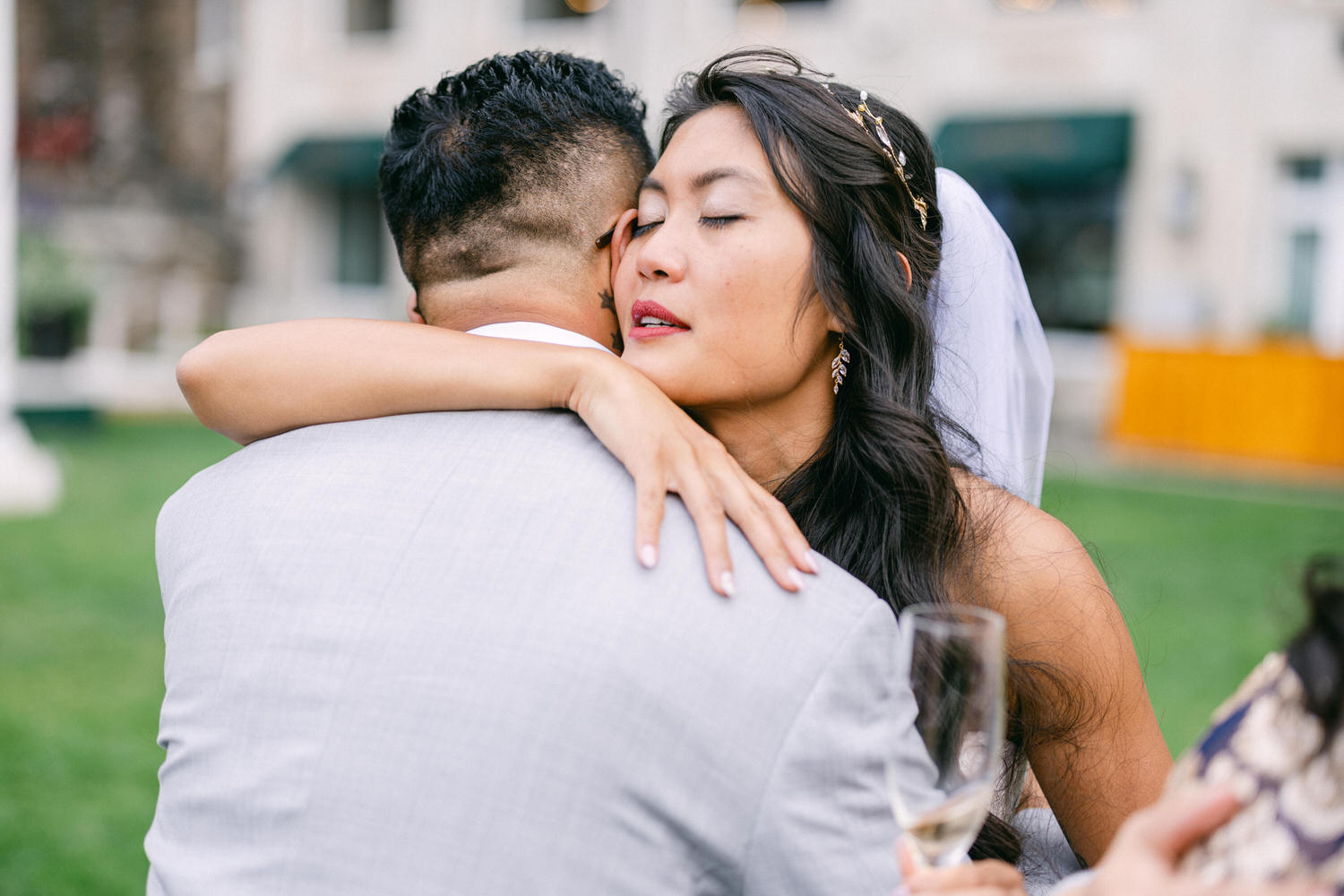 A bride shares a heartfelt moment with her groom, embracing him while wearing a beautiful wedding gown and veil, set against a lush green background.