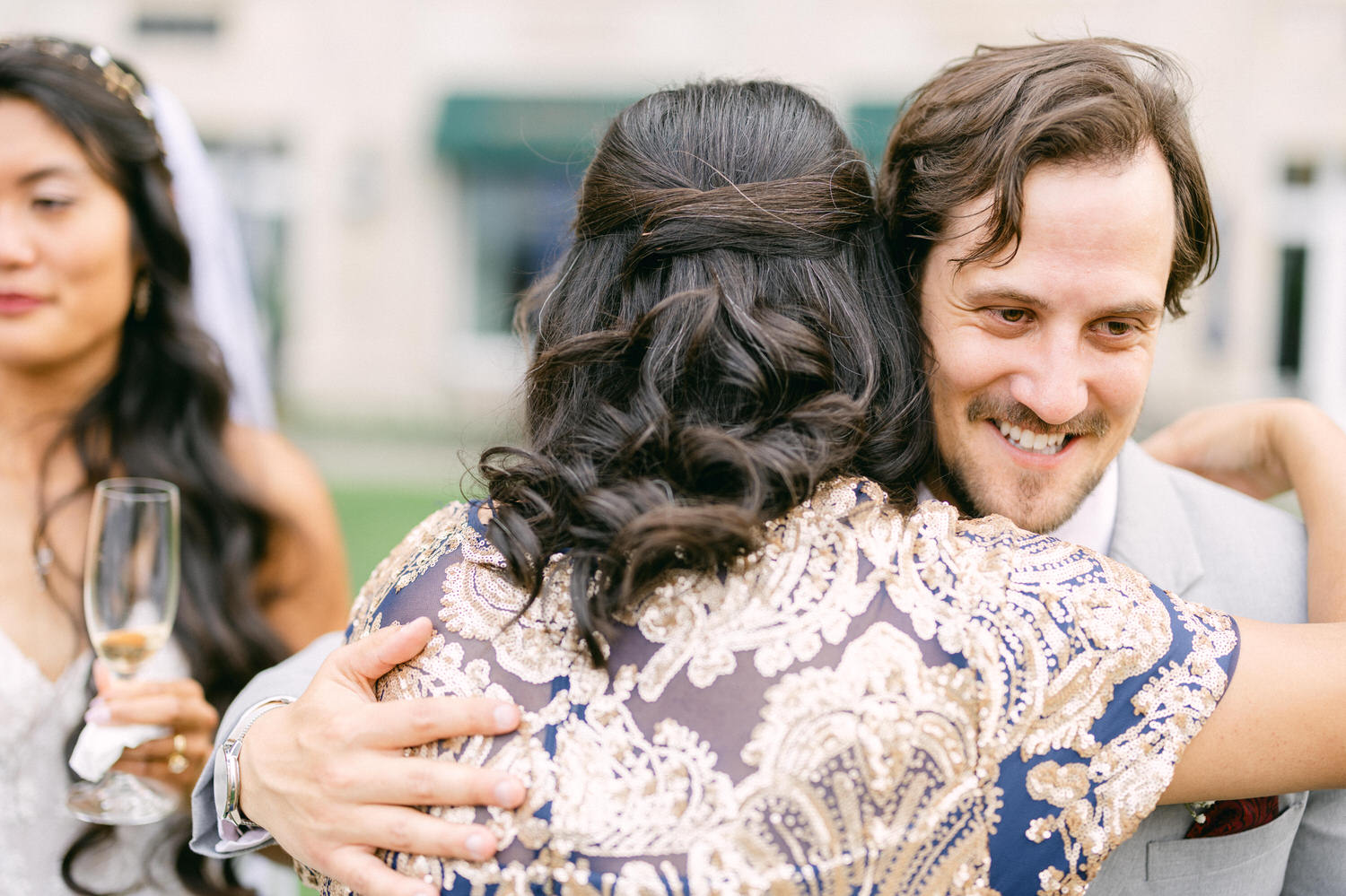 A man smiles warmly as he embraces a woman in a beautiful, embellished dress, with another woman in the background holding a champagne glass.