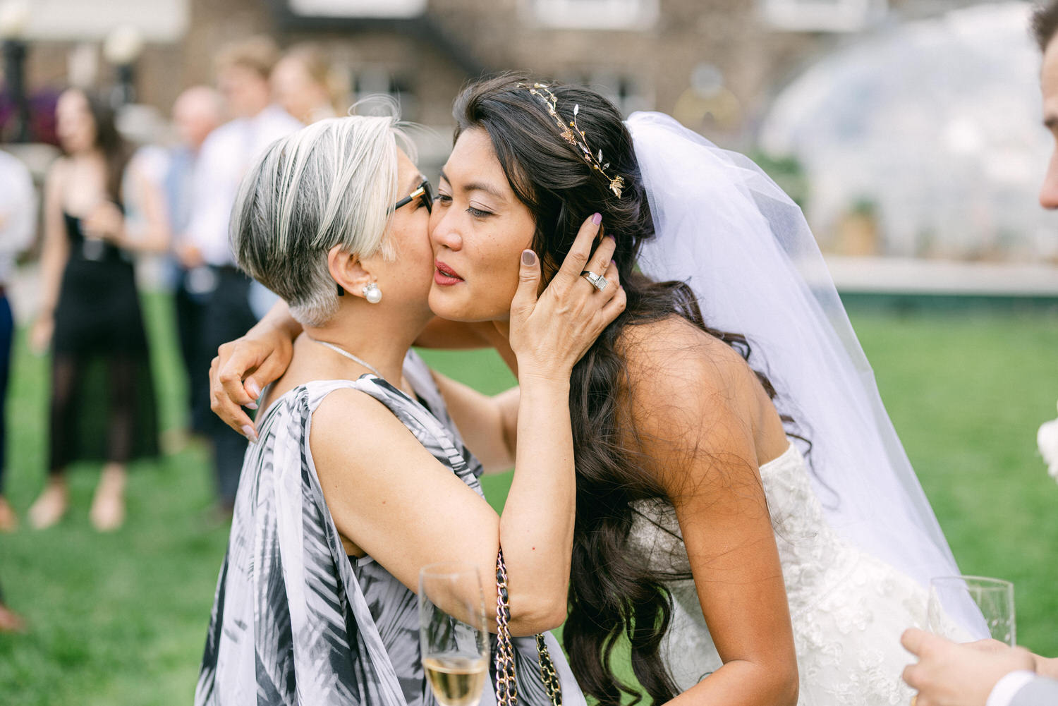 A bride and her mother share a heartfelt embrace at an outdoor wedding celebration, with guests in the background.