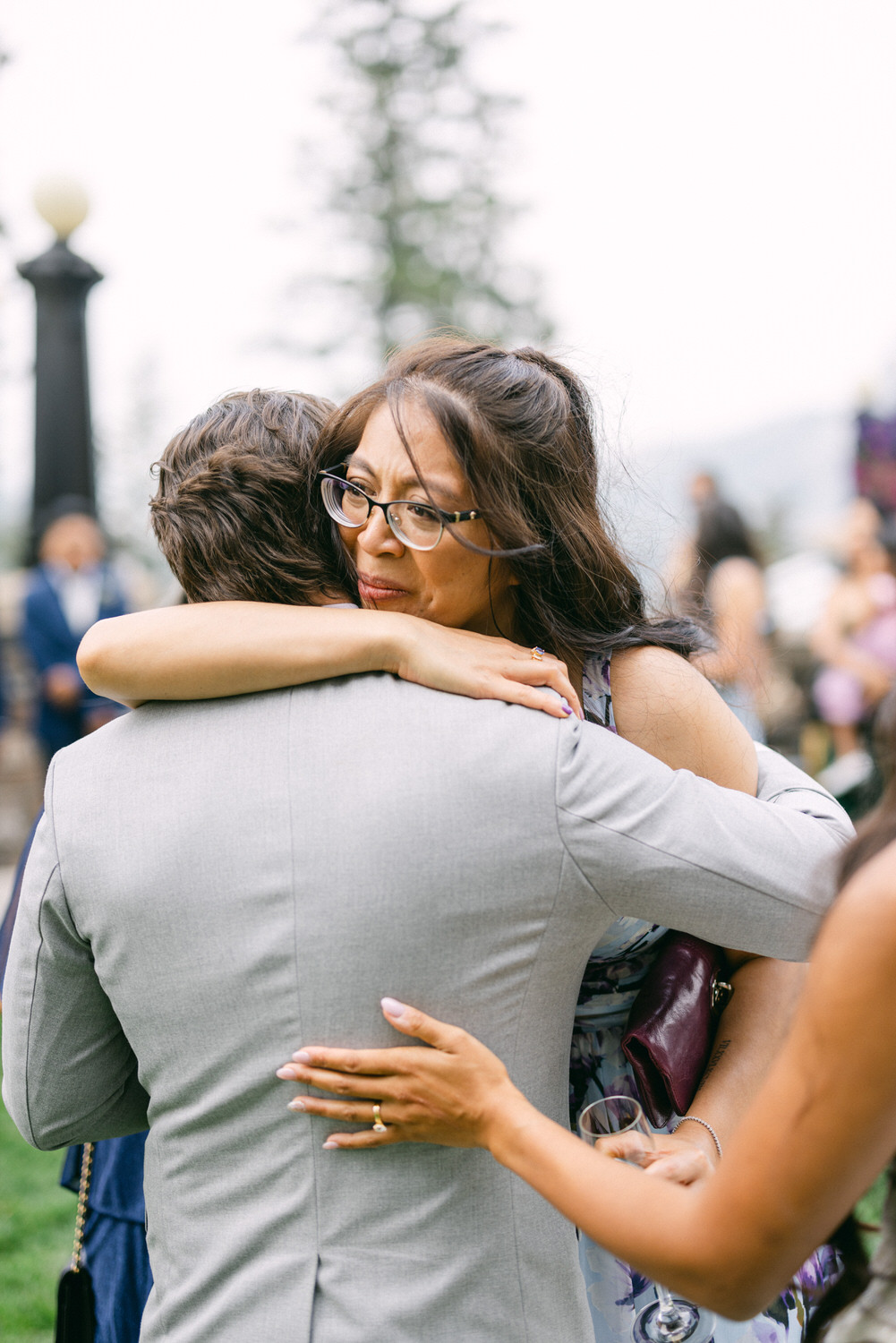 A woman embraces a man in a heartfelt moment, expressing deep emotions at a social gathering, surrounded by blurred guests and greenery in the background.