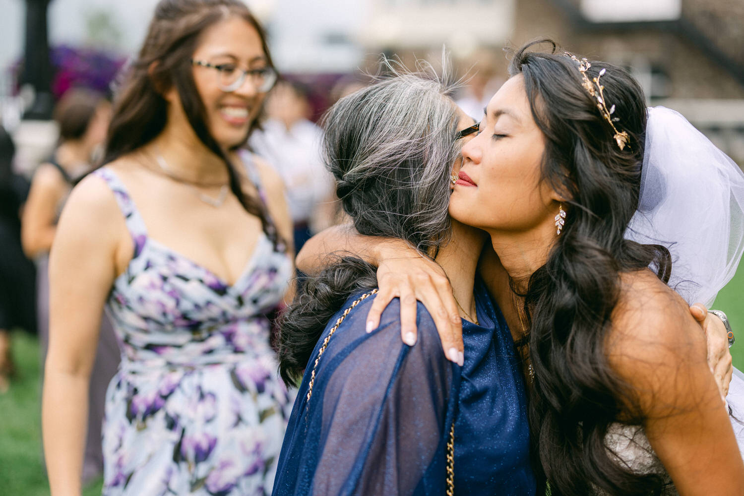 A bride shares a heartfelt hug with her mother while a smiling guest looks on in the background, celebrating a joyful moment at a wedding.