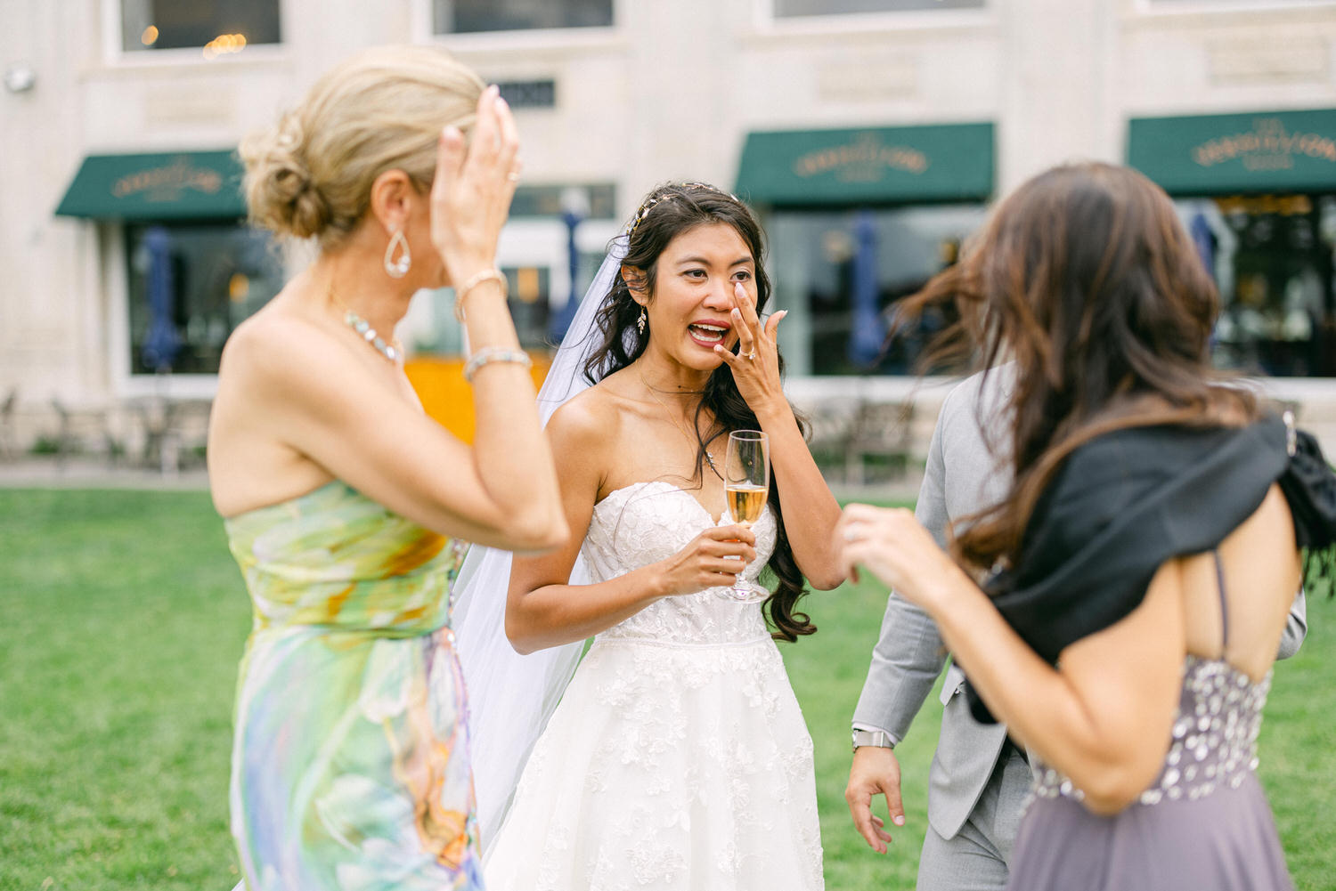 A bride, emotionally overwhelmed, wipes her tears while holding a champagne flute amidst joyful friends during a wedding celebration.