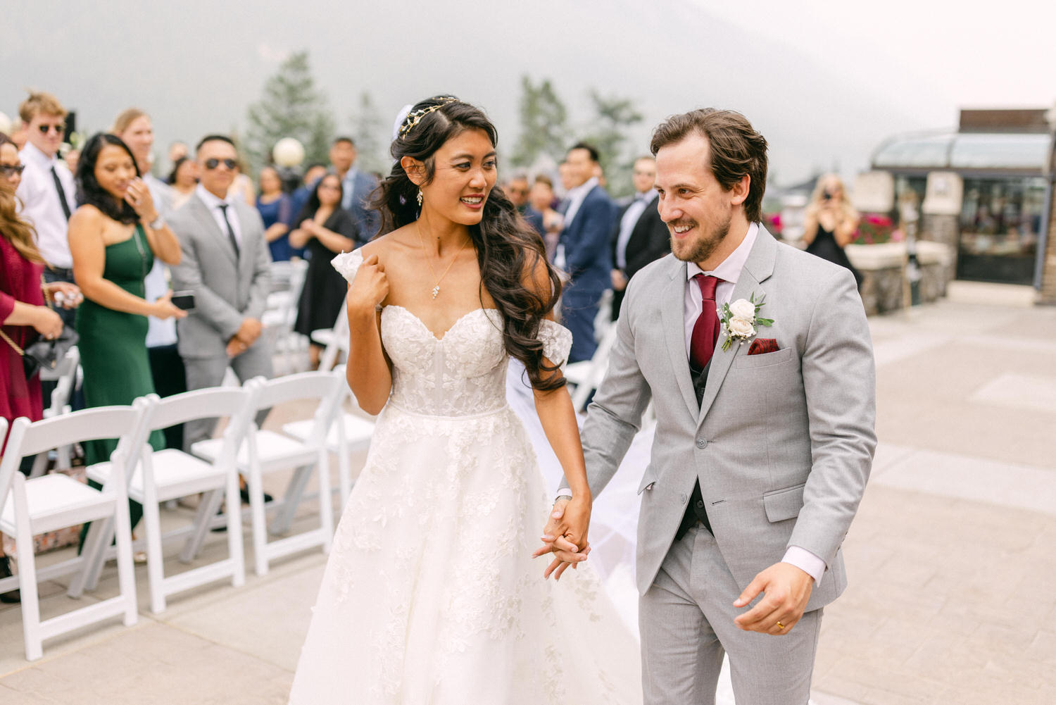 A bride and groom smile and hold hands as they walk down the aisle, surrounded by guests in a beautiful outdoor setting.