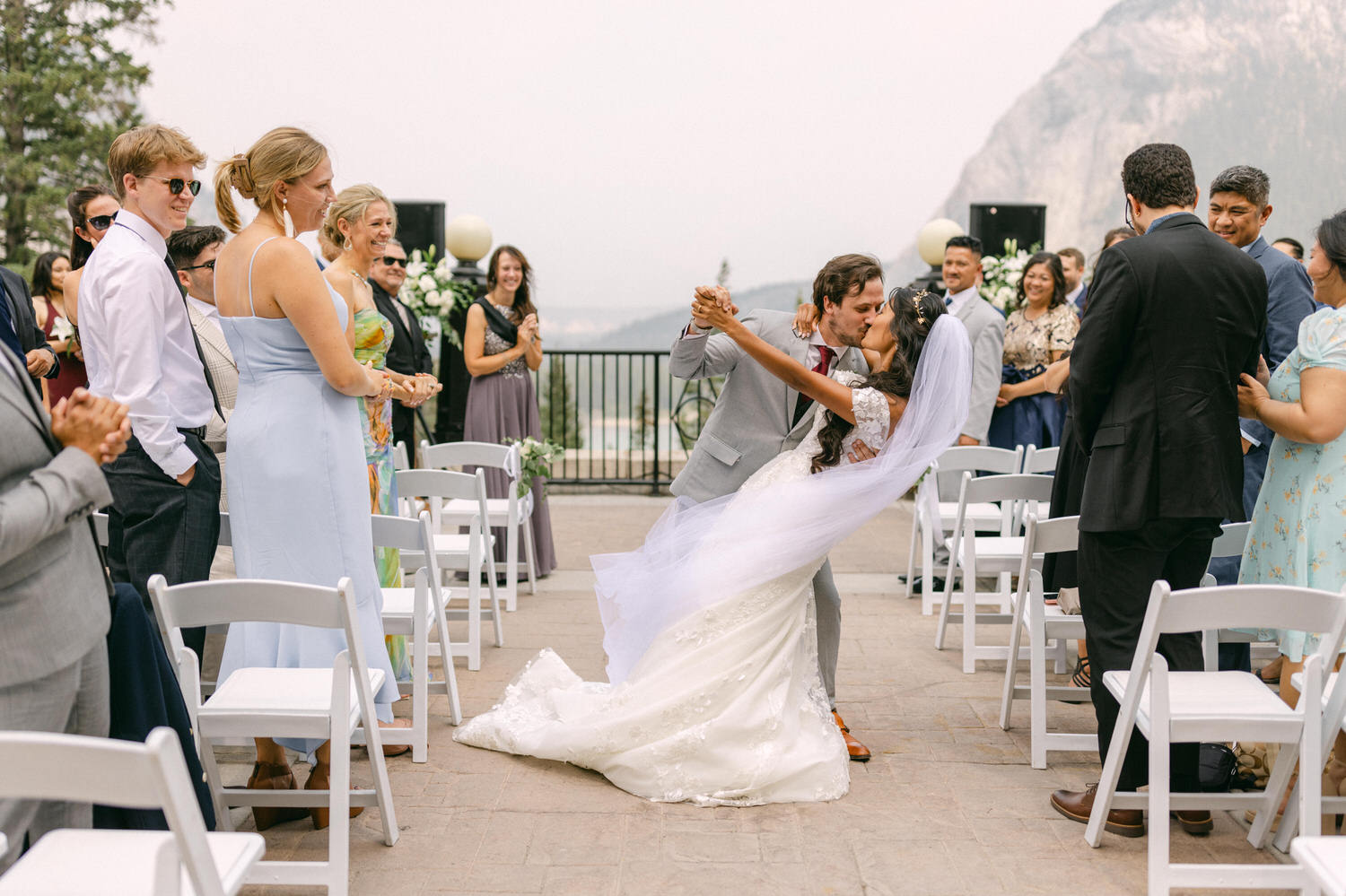 A joyful moment as the bride and groom share a kiss during their outdoor wedding ceremony, surrounded by guests and scenic mountains.