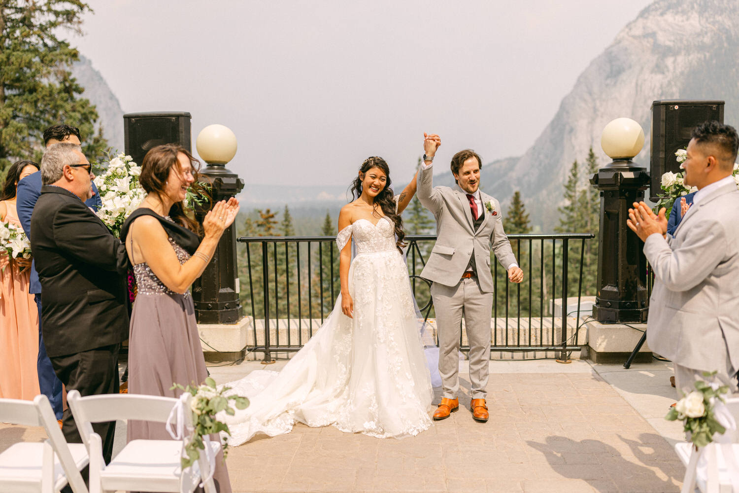 A joyous couple celebrates their marriage outdoors with a picturesque mountain backdrop, surrounded by applauding guests.