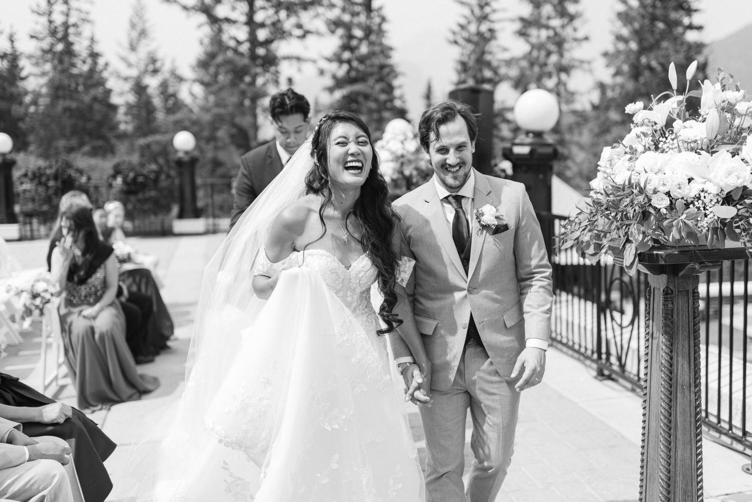 A bride and groom laughing and holding hands as they exit their wedding ceremony, surrounded by guests and floral decorations.