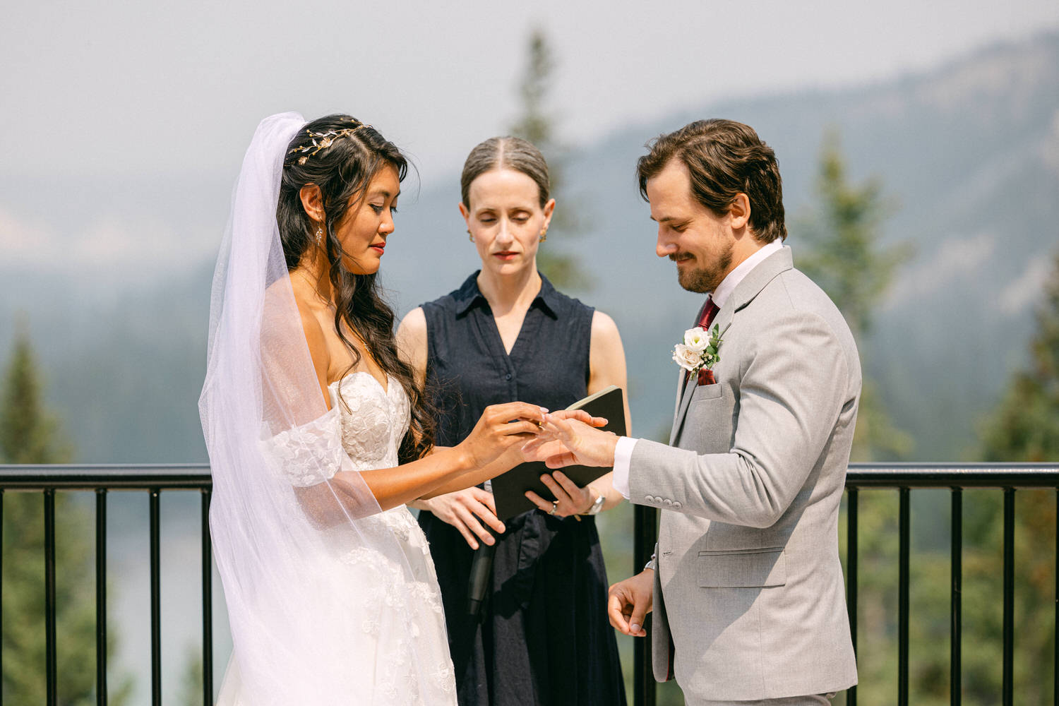 A bride and groom exchange rings during their outdoor wedding ceremony, with an officiant present and a scenic mountain backdrop.