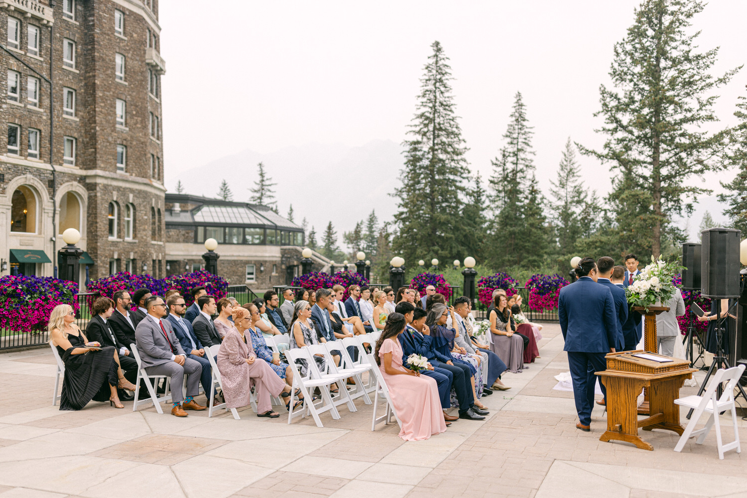 Guests seated in elegant attire at a wedding ceremony with a charming building and floral arrangements in the background.