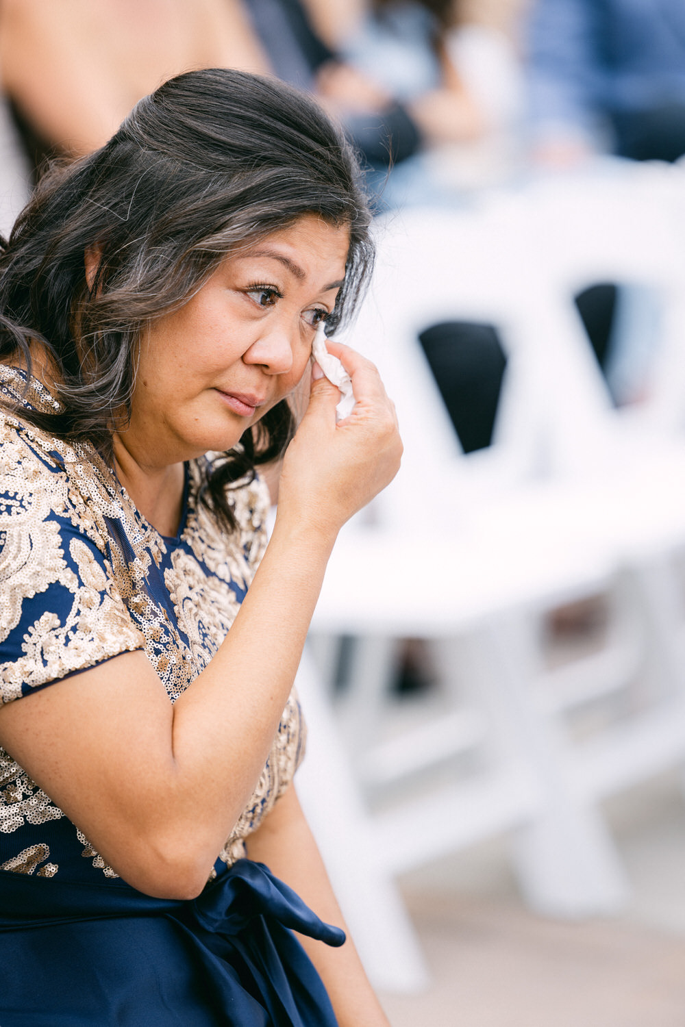 A woman with long dark hair wearing a sequined dress wipes away tears during a heartfelt moment at an event.