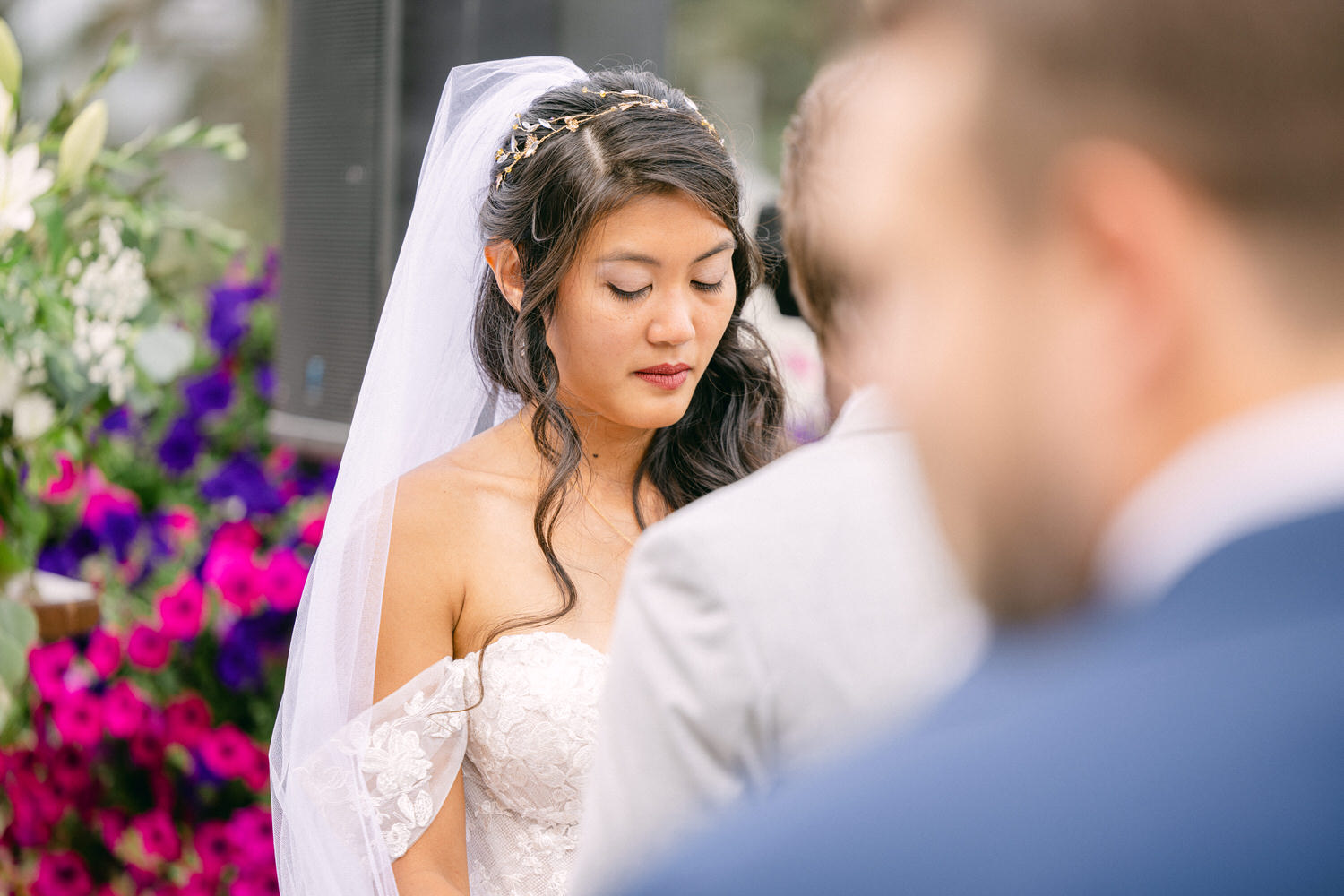 A bride with her eyes closed during a wedding ceremony, surrounded by flowers and guests.