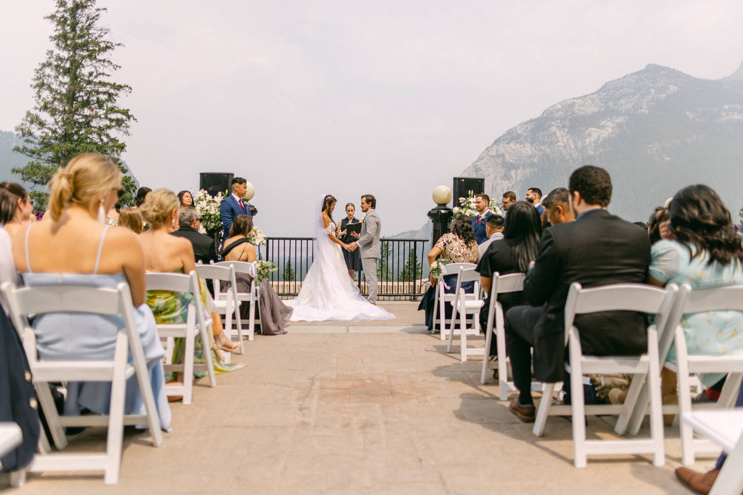 A couple exchanges vows during an outdoor wedding ceremony, surrounded by guests and scenic mountains in the background.