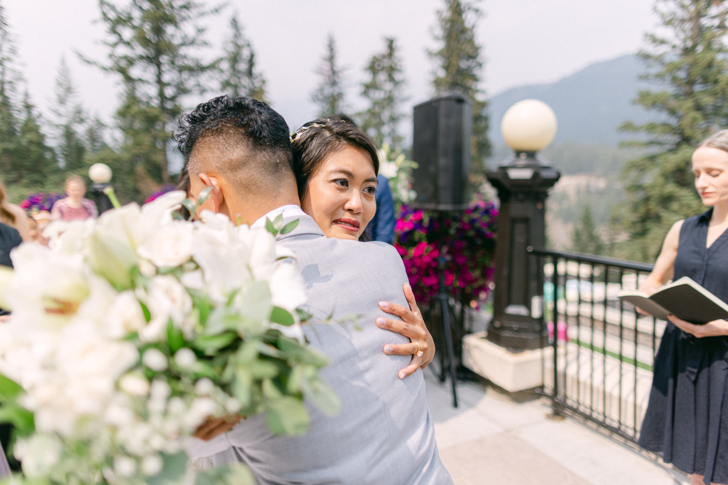 A couple shares a heartfelt hug during their wedding ceremony, surrounded by floral decorations and guests in a scenic outdoor setting.