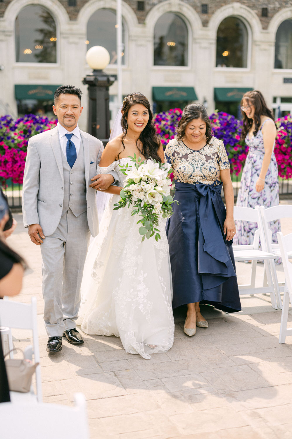 A bride in a flowing white gown, holding a bouquet, smiles as she walks arm-in-arm with her father and mother towards a wedding ceremony, surrounded by vibrant floral decorations.