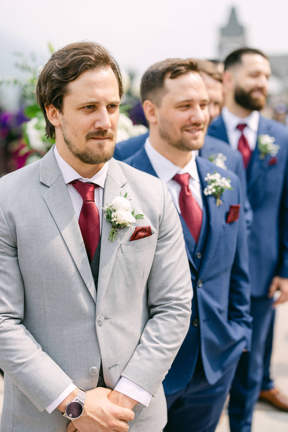 A group of well-dressed men, including the groom in a grey suit and red tie, standing together at an outdoor wedding ceremony with floral decorations in the background.