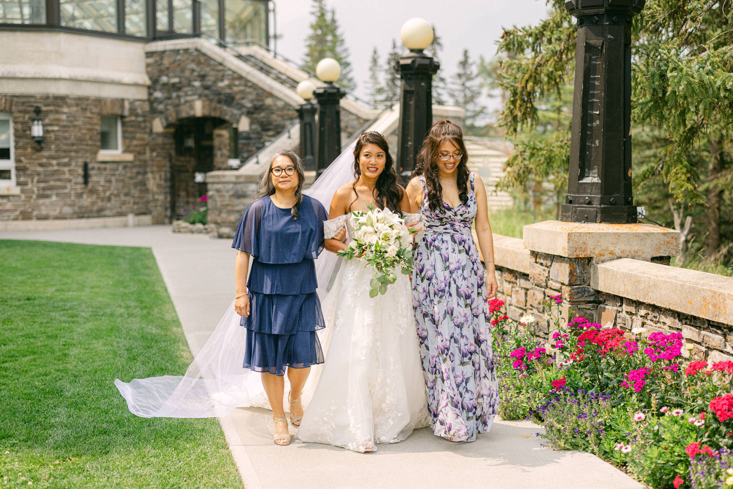 A bride in a stunning white gown is joyfully walking with two friends, one in a navy dress and the other in a floral gown, surrounded by lush greenery and colorful flowers.