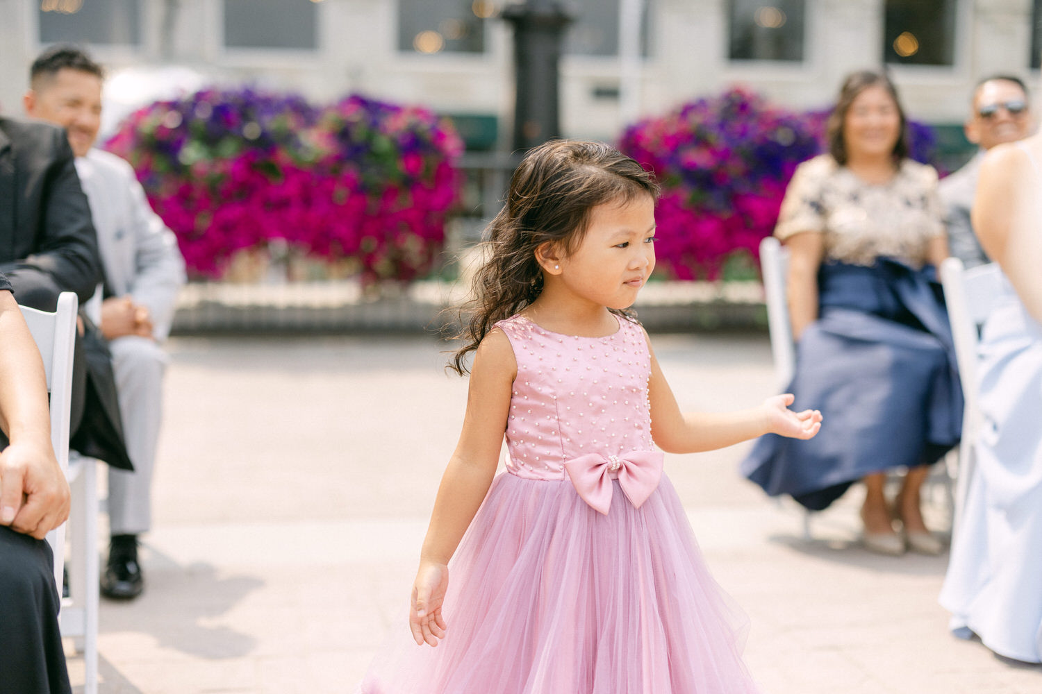 A young girl in a pink dress with a bow walks gracefully during a wedding ceremony, with guests seated in the background and vibrant purple flowers decorating the scene.