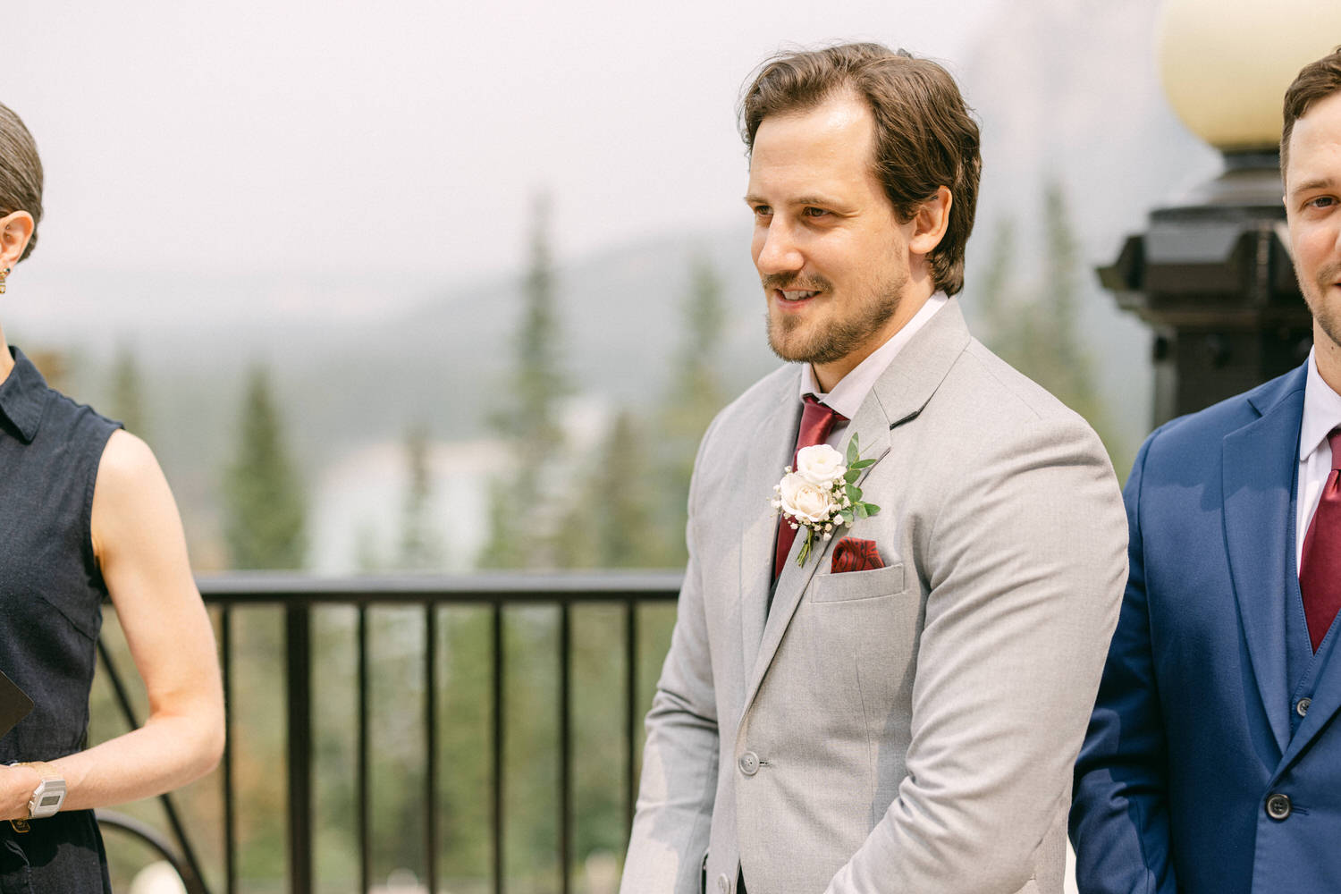 A man in a light gray suit with a maroon tie and boutonniere stands smiling during an outdoor wedding ceremony, accompanied by a woman in a dark dress and another man in a blue suit.