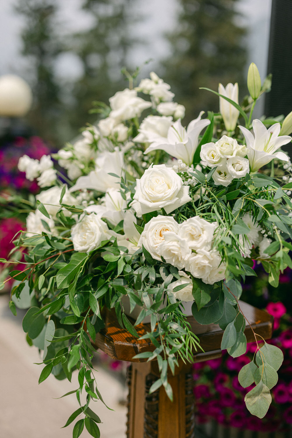 A close-up of a lush floral arrangement featuring white roses and lilies, accented with greenery, placed on a wooden pedestal, with colorful flowers in the background.